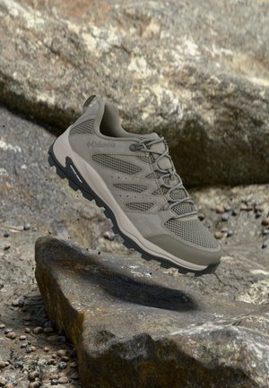 Trailrunningschoen met mesh, suède panelen en een robuuste zolen die zweeft boven rotsachtig terrein, ontworpen voor buitenactiviteiten.