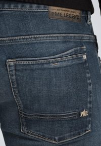 Jeans en denim de couleur bleu foncé avec une finition texturée. Comprend une étiquette en cuir sur la taille et une poche arrière avec des détails de couture.