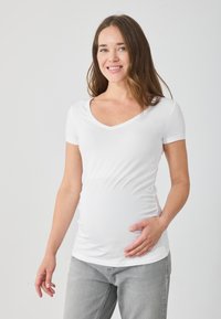 Balta grūtniecības T-kreklu ar īsām piedurknēm un V-veida kakla izgriezumu, ar pieguļošu dizainu un sasējušām malām. Apvienots ar pelēkiem džinsiem.