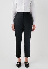 Zwarte maatwerkbroek met een slim fit, platte voorkant en subtiele glans. Gepaard met een wit overhemd met lange mouwen en zwarte instappers.