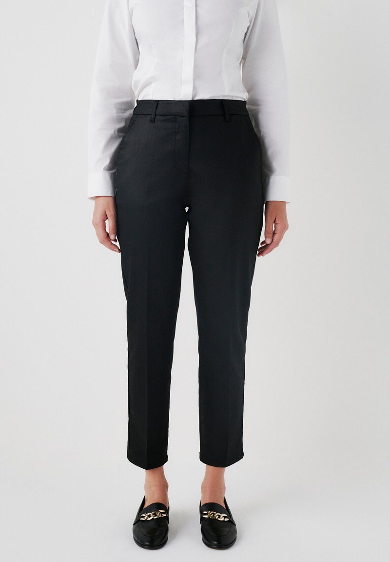 Zwarte maatwerkbroek met een slim fit, platte voorkant en subtiele glans. Gepaard met een wit overhemd met lange mouwen en zwarte instappers.