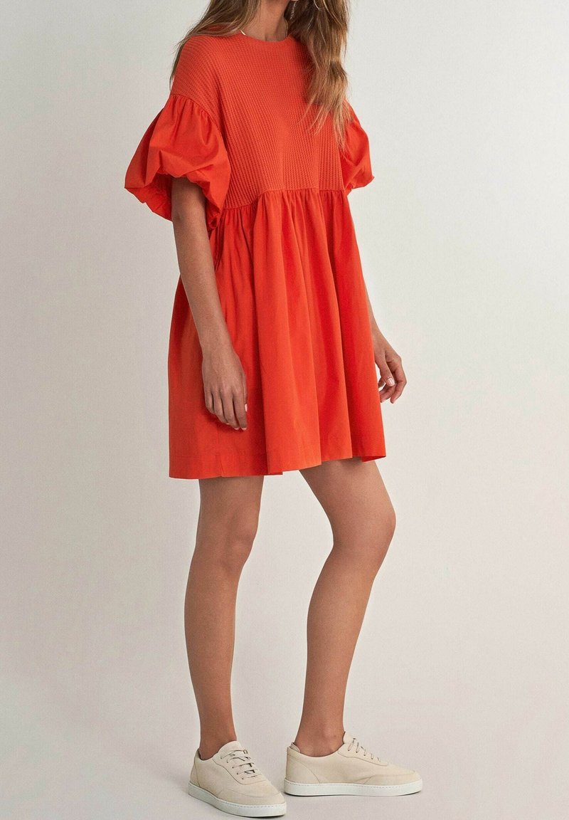 Vestido corto rojo de manga corta con un cuerpo texturizado, falda fruncida y mangas abullonadas elásticas. Combinado con zapatillas de color claro.