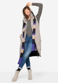 Graue ärmellose Steppweste mit Kapuze, kombiniert mit einem gestreiften Langarmshirt, blauen Jeans und beigen Stiefeletten. Gefranster Schal mit Streifen.