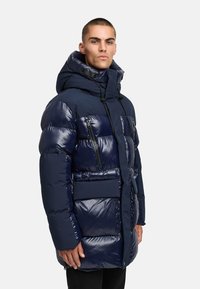 Meripuku takki kiiltävä pinta, suuri huppu, vetoketjulliset rintataskut ja leveä vyötärö. Ominaisuuksiin kuuluu moderni, laatikkomainen siluetti.