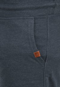 Un pantalon de survêtement gris foncé avec une taille côtelée, une poche avant et un petit patch en cuir marron près de la poche pour le détail.