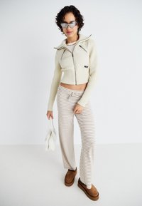 Cropped cream zip-up jacket with ribbed texture, striped beige wide-leg pants, brown slip-on shoes, and white handbag. Reflective sunglasses.