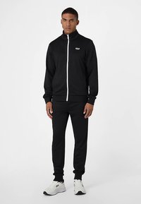 Sort træningsjakke med hvid lynlås og logo, matchende sorte bukser. Stoffet er glat med en tætsiddende pasform. Bruges med hvide sportssko.