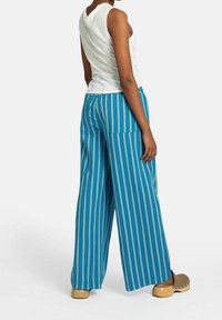 Kvinna i blåa och gröna vertikalt randiga vida byxor, vit ärmlös topp och beige träskor med träklackar, stående på sidan.