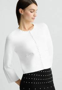 Cardigan bianco testurizzato con dettagli in argento lungo il collo e le tasche, abbinato a una gonna nera con decorazioni in argento.