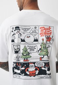 Balts kokvilnas T-krekls ar komiksu stila grafiku, kurā redzami sniegavīri, Ziemassvētku eglītes un Ziemassvētku vecītis. Teksta elementi ietver "Deck the Halls" un runas burbuļus.
