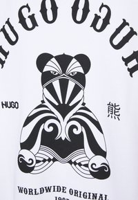 Ours stylisé en noir et blanc avec des motifs tribaux, encadré par le texte « HUGO », au-dessus de « WORLDWIDE ORIGINAL 1987 » sur fond blanc.