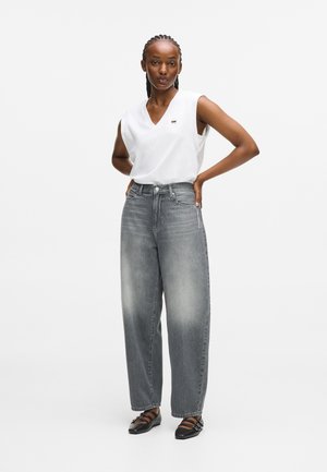 Witte mouwloze top met een v-hals, gecombineerd met hoge, wijde grijze jeans met subtiele vervaging. Zwarte platte schoenen met banden.