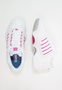 Ett par vita sportskor med rosa ränder, där den ena visar en ovanifrån vy med snören och varumärkesnamn, medan den andra visar sidovy och detalj av sulan.