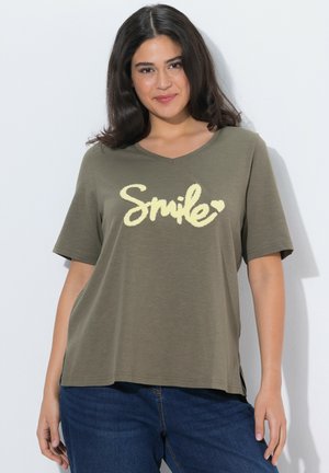 Giovane donna con capelli scuri sorride, indossa una maglietta verde oliva con la scritta "Smile" e jeans blu su sfondo bianco.