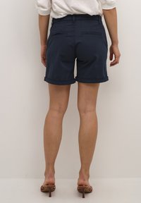 Tumed sinise värvi puuvillased lühikesed püksid, millel on rullitud äär, kahte taga- Taskut ja sile tekstuur, kantud pruunide kontsaga sandaalidega.