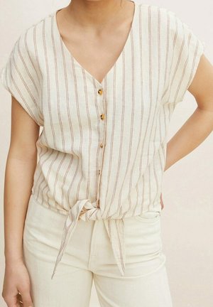 Femme portant un chemisier rayé couleur crème à manches courtes, boutonné, noué à la taille, avec un pantalon clair, mains posées sur les hanches.