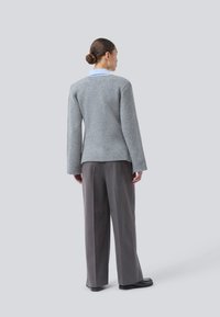 Kvinde set fra ryggen iført en grå sweater over en blå skjorte med krave, løse mørkegrå bukser og sorte sko, mod en ensfarvet baggrund.