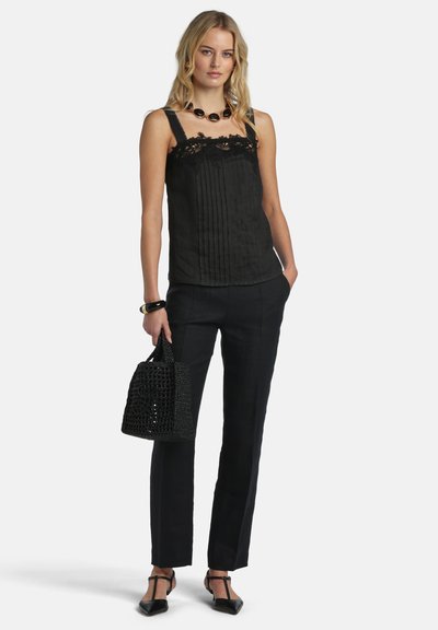 Femme portant un haut sans manches noir orné de dentelle, un pantalon noir, des talons noirs à bride en T, tenant un sac à main noir tissé, debout devant un fond blanc.