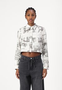 Hvit avkortet skjorte med et diskret svart abstrakt mønster, med krage og lange ermer, kombinert med høyere svarte jeans.
