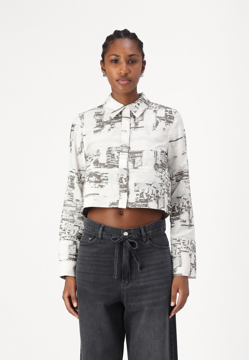 Hvit avkortet skjorte med et diskret svart abstrakt mønster, med krage og lange ermer, kombinert med høyere svarte jeans.