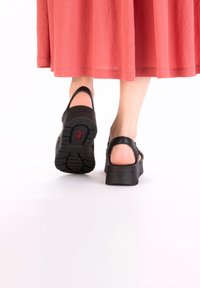 Sabots noirs avec une épaisse plateforme, une sangle arrière ajustable et une semelle texturée en caoutchouc. Portés avec une jupe midi corail, mettant en valeur le design du talon.