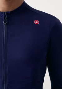 Marineblaue Fahrradjacke aus strukturiertem Stoff, mit durchgehendem Reißverschluss und einem runden roten Logo auf der linken Brustseite. Figurbetontes Design, lange Ärmel.