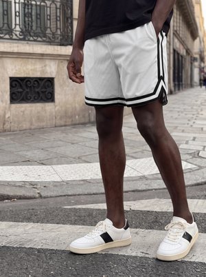 Mand iført hvide shorts med sorte striber og hvide sneakers med sorte detaljer, der står på et fodgængerfelt i en by.