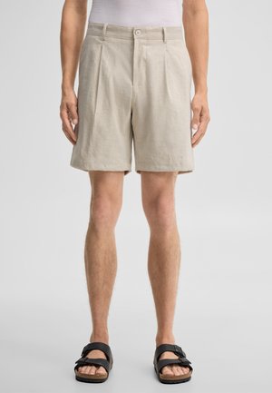 Mann trägt beige plissierte Shorts, weißes Hemd eingesteckt und schwarze Sandalen mit Schnalle, steht vor einfachem Hintergrund.