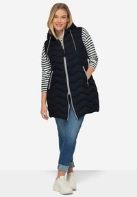 Marinblå quiltad puffväst med huva, med en framzipper och sidofickor, tillsammans med en randig långärmad topp och denimjeans.