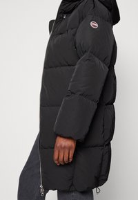 Black puffer jacket with a hood, featuring a zip closure, oversized quilted design, and logo patch on the left arm. Smooth fabric texture.