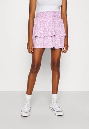 Vrouw die een wit tanktop draagt, een roze bloemenprint gelaagde minirok, witte sokken en witte hoge sneakers, staand tegen een effen witte muur.