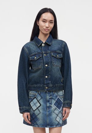 Femme aux longs cheveux foncés portant une veste en jean bleue et une jupe en jean assortie avec des patchs losanges à carreaux, debout devant un fond uni.