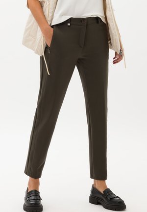 Personne portant un pantalon slim couleur olive foncé, des mocassins noirs épais, un haut blanc et une veste sans manches beige, les mains dans les poches.