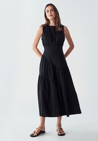 Črna brezrokavnica z oprijetim topom, slojevitim robom in diagonalnimi šivi. Narejena je iz mehkega blaga; model je stiliziran z črnimi sandali.