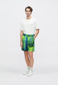 Bunte Badeshorts mit einem lebhaften grünen und blauen abstrakten Muster, kombiniert mit einem weißen T-Shirt und weißen Turnschuhen.