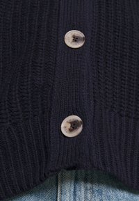 Námořnický pletený kardigán s ribbed texturou a dvěma kulatými, světlými knoflíky s logem. Spodní část je vyrobena z denimu.