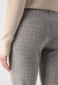Gros plan d'une personne portant un pantalon gris à motif chevrons et un pull en maille beige, main reposant près de la cuisse.
