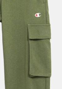 Ελαφρώς ελαιώδεις πράσινες υφασμάτινες παντελόνες με μεγάλη πλευρική τσέπη που έχει καπάκι και μικρό λογότυπο της μάρκας Champion πάνω από την τσέπη.