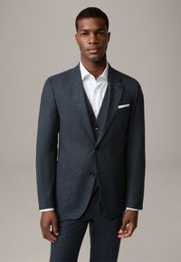 Completo grigio scuro con una giacca a rever a punte, due tasche anteriori e un gilet abbinato. Camicia bianca con colletto classico.