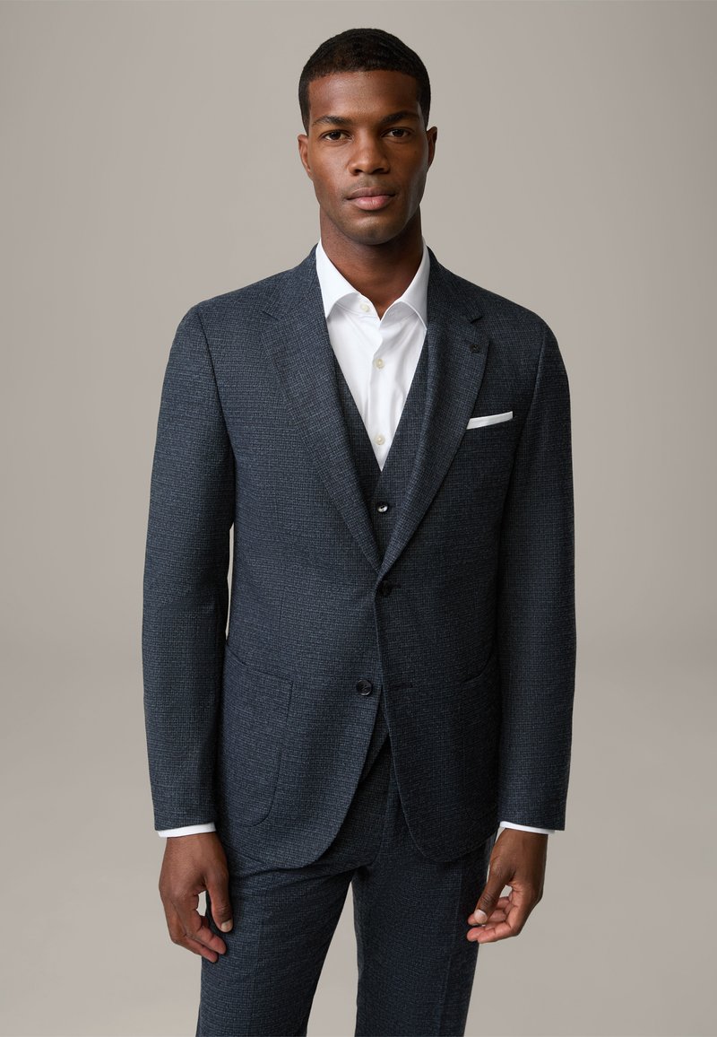 Completo grigio scuro con una giacca a rever a punte, due tasche anteriori e un gilet abbinato. Camicia bianca con colletto classico.