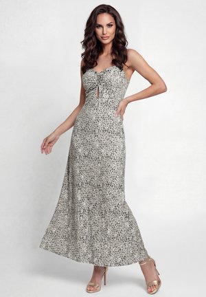 Nő áll egy ujjatlan, bokáig érő, szürke-fehér állatmintás ruhában, melynek mellrésze ráncolt, és aranyszínű magassarkú szandált visel, egy egyszerű háttér előtt.