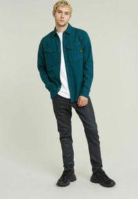 Camicia verde acqua con bottoni e due tasche sul petto, indossata sopra una t-shirt bianca, abbinata a pantaloni slim-fit scuri e sneaker nere.