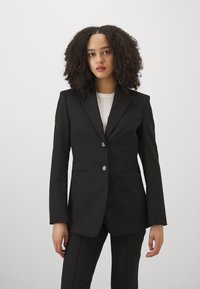 Young woman with curly hair wearing a fitted black blazer over a white top, standing against a plain light background.