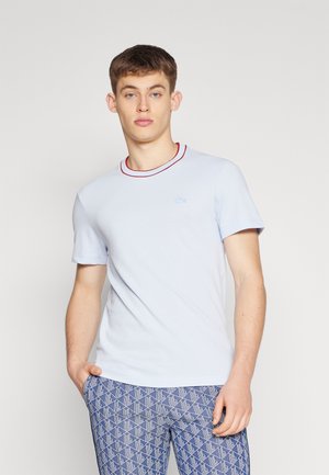 Camiseta de algodón azul claro con cuello de canalé rojo, que presenta un pequeño logotipo bordado en el lado izquierdo del pecho. El modelo lleva pantalones con estampado.