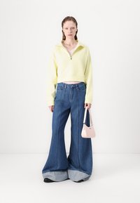 Gelbes, verkürztes Sweatshirt mit Viertelreißverschluss, kombiniert mit weit geschnittenen blauen Jeans mit einem kontrastierenden Denim-Saum und einer kleinen pinken Handtasche.