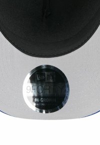 Schwarze Kappe mit einem grauen Schirm, ausgestattet mit einem runden metallischen Etikett mit der Aufschrift "9FORTY M-CROWN ADJUSTABLE." Strukturierter Stoff mit blauen Akzenten.