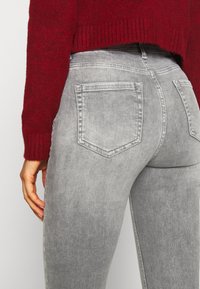Jeans de mezclilla gris con un diseño ajustado, que cuentan con dos bolsillos traseros y una textura ligeramente descolorida, combinados con un suéter de punto rojo.