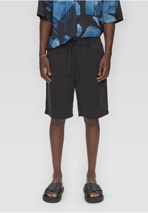 Man draagt zwarte shorts met trekkoorden, zwarte slides en een shirt met korte mouwen met een blauw-zwart abstract patroon, staand tegen een grijze achtergrond.