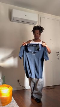 Muškarac s dredovima u bijeloj majici bez rukava drži plavu polo majicu u zatvorenom prostoru, blizu vrata i klima uređaja na bež zidu.