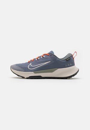 Niebiesko-szary but do biegania z białą podeszwą, pomarańczowymi sznurowadłami, białym logo Nike i czerwoną pętlą na pięcie, pokazany z profilu na białym tle.
